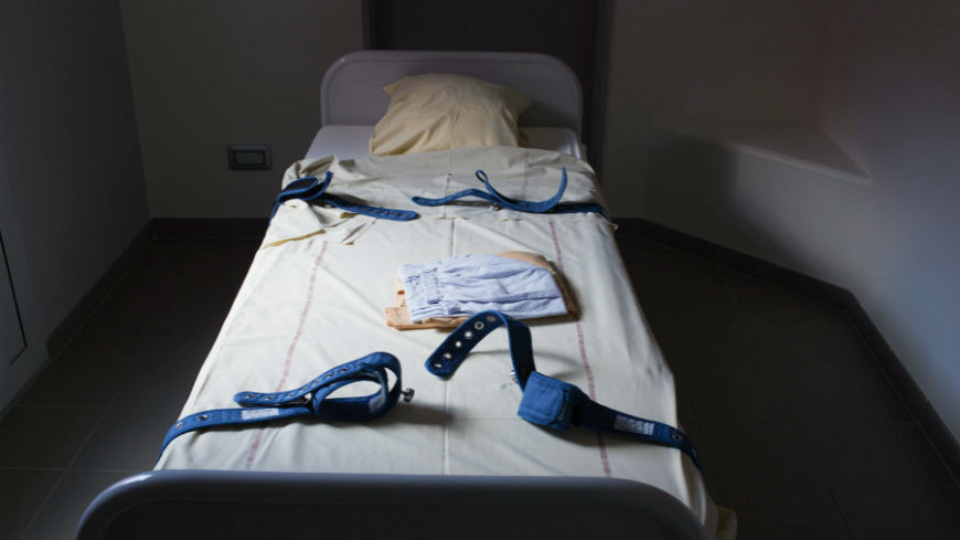 Exemple d'une chambre d'isolement dans laquelle il n'y a qu'un lit de contention. Sur ce lit blanc sont fixées 4 sangles ajustables en tissu bleu. Un pyjama d'hospitalisation ainsi qu'un léger drap sont pliés au centre du matelas. Le lit a été fait. La chambre n'est éclairée que par la lumière naturelle. Les murs sont blancs et le sol est gris.