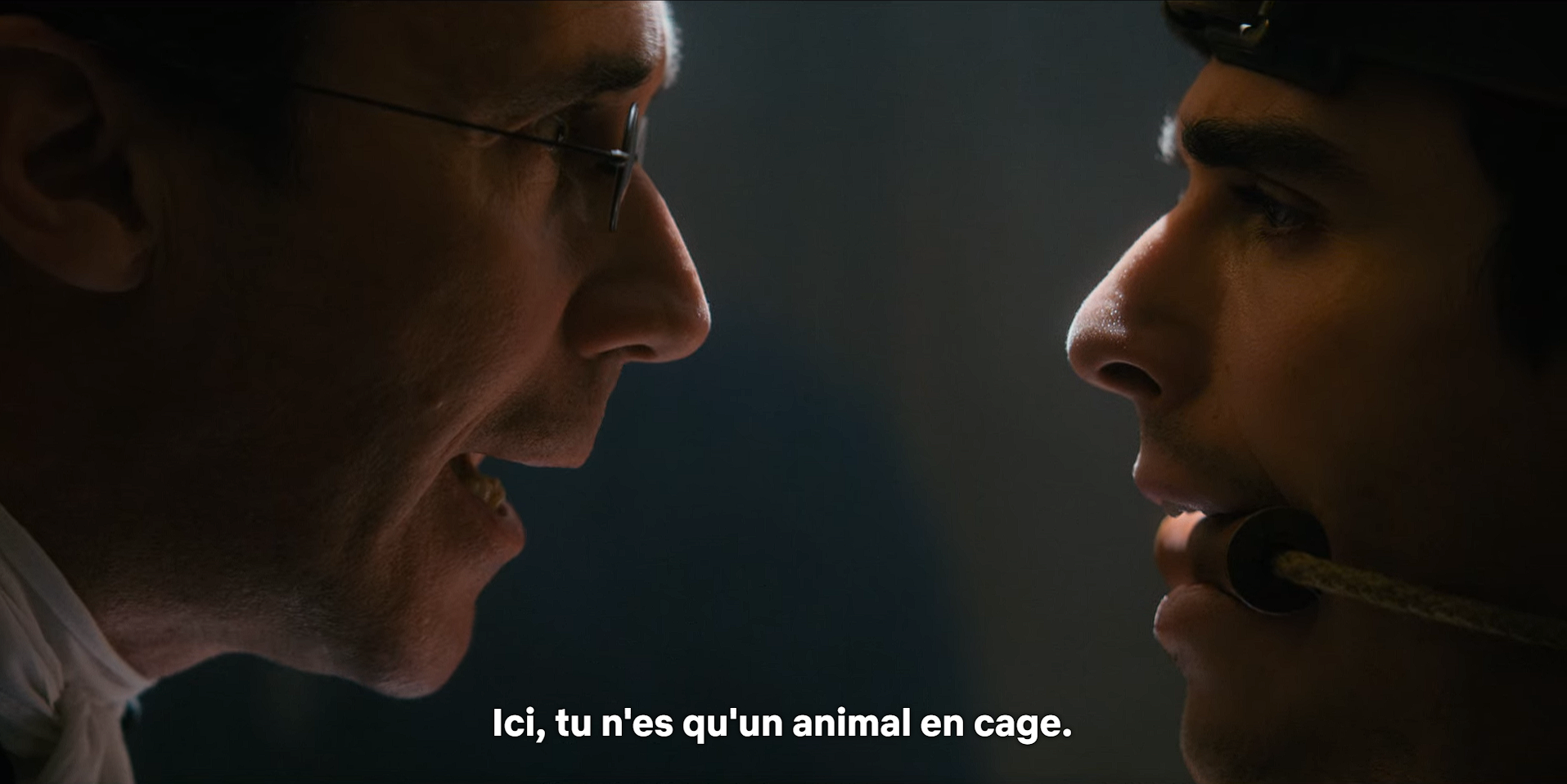 Capture d'écran de la série La Reine Charlotte, épisode 4, scène où le docteur monro prend le roi george les yeux dans les yeux et dit "ici tu n'es qu'un animal en cage". Le plan est très près de leurs visages et coupe leurs fronts. Le roi george est assis et baillonné. Il regarde le docteur avec crainte.